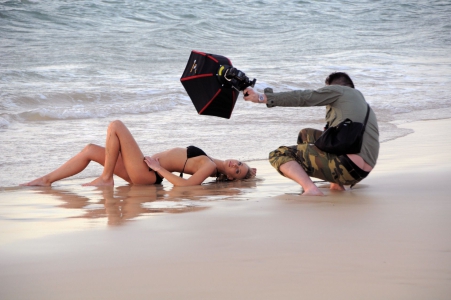 Eine Frau liegt am Wasserrand eines Strandes, während ein Mann mit einer Kameraausrüstung kniet und sie filmt.