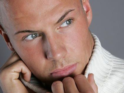 Nahaufnahme eines jungen, blonden Mannes mit intensivem Blick, der nachdenklich in die Ferne blickt und einen hellen Pullover trägt.