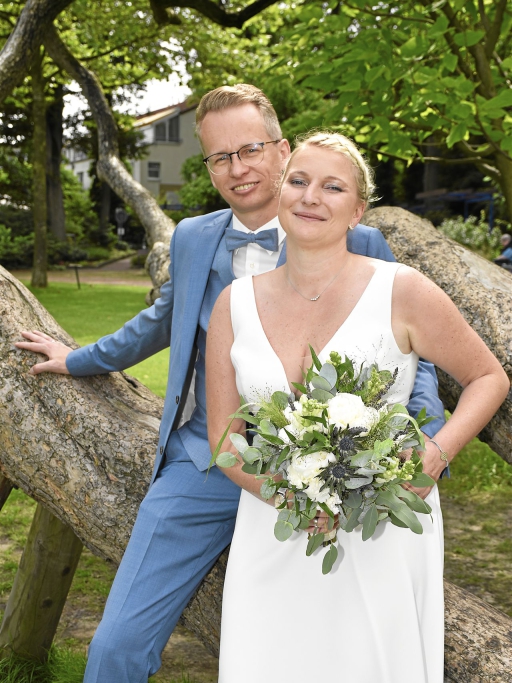 Ein Paar posiert vor einem großen Baumstamm in einem parkähnlichen Ambiente, die Frau hält einen Blumenstrauß.