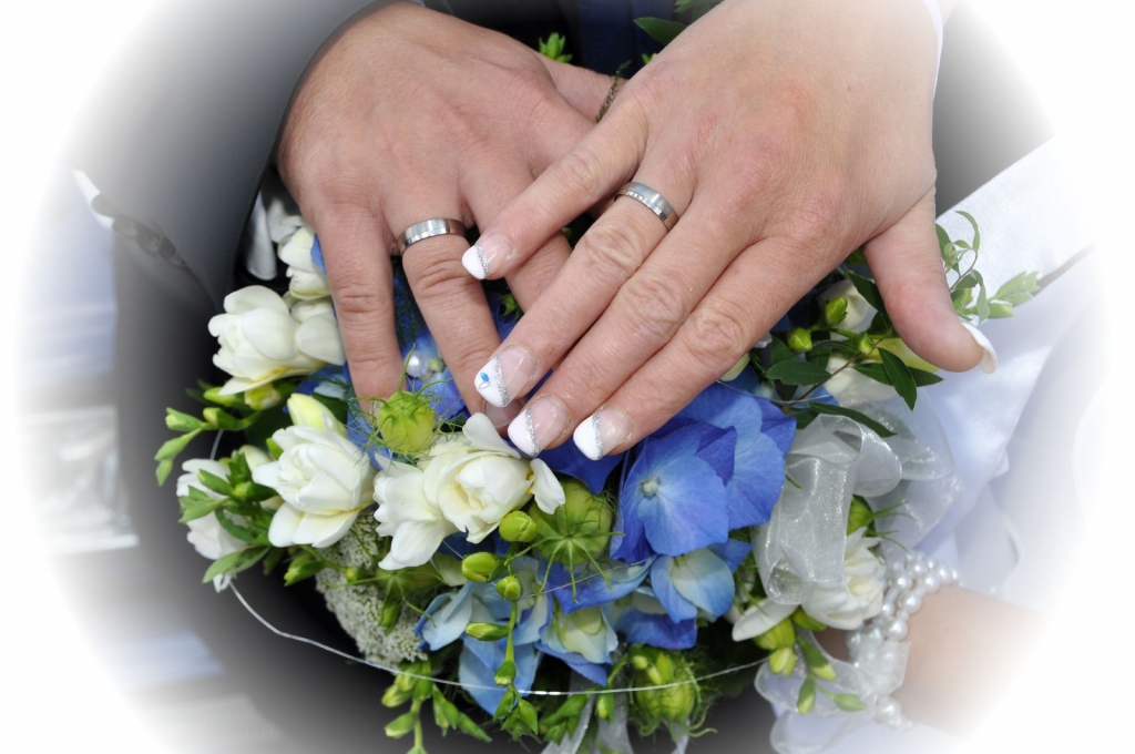 Zwei Hände mit mehreren Ringen ruhen auf einem Blumenstrauß aus weißen und blauen Blüten.