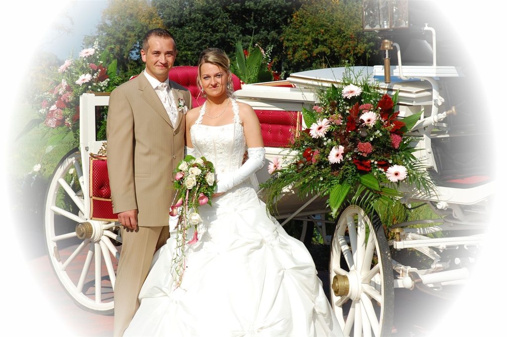 Ein Brautpaar posiert vor einem dekorativen Hochzeitswagen, der reich mit Blumen geschmückt ist.